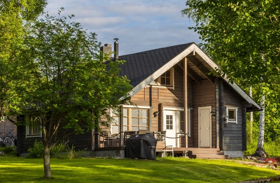 Brownish log cottage in the summer setting, with green grass in the front and leafy trees.